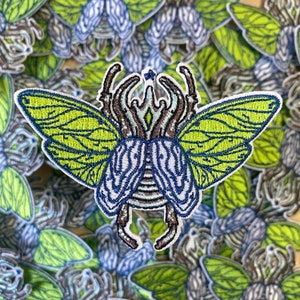 May include: An embroidered patch depicting a beetle with wings, featuring a detailed design in green and blue. The wings and body are adorned with intricate patterns.