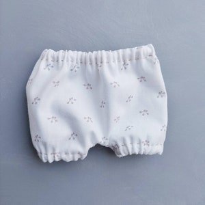 May include: White cotton baby bloomers with a small floral print. The bloomers have a gathered elastic waistband and are made of soft, breathable fabric.