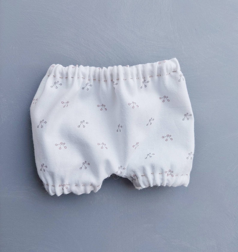 May include: White cotton baby bloomers with a small floral print. The bloomers have a gathered elastic waistband and are made of soft, breathable fabric.