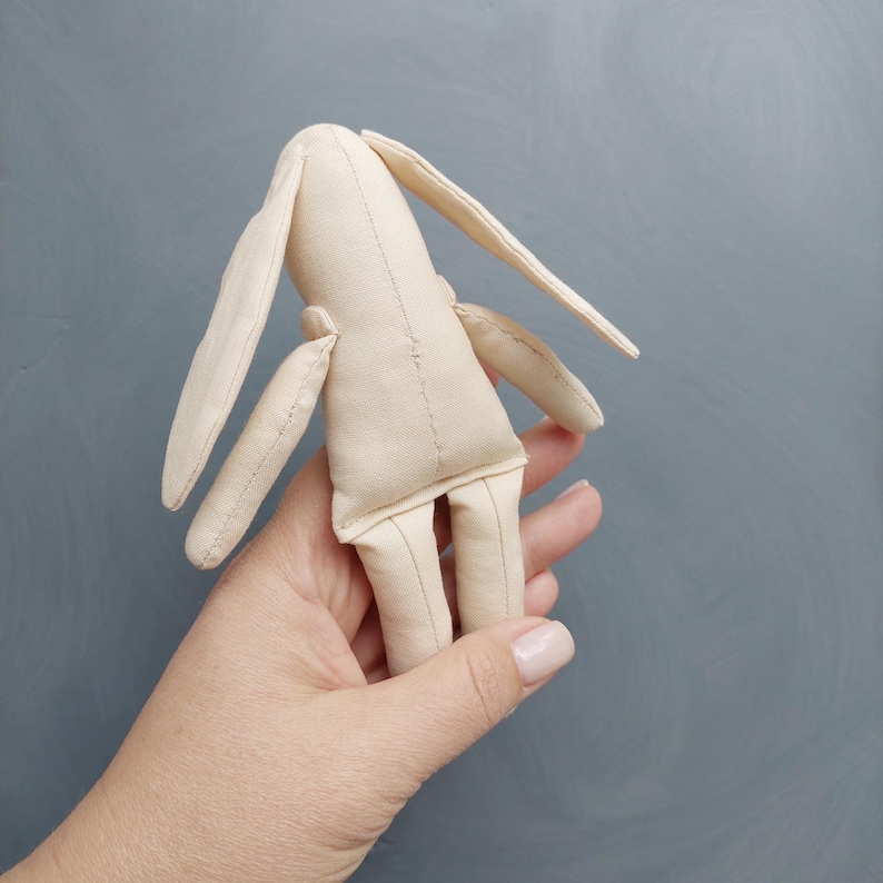 May include: A beige fabric bunny toy with long ears and stitched details. The toy is held in a hand.