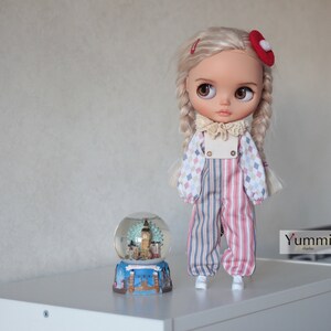 May include: A doll with blonde braids wearing a red and blue striped jumpsuit with a white collar and a red bow on her hair. The doll is standing next to a snow globe with a London skyline inside.