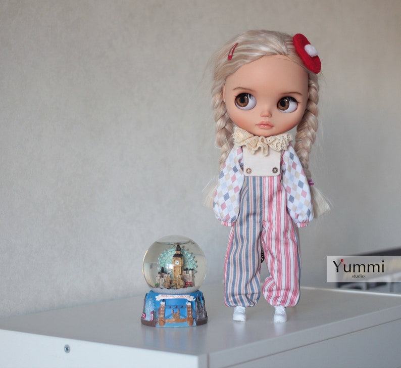 May include: A doll with blonde braids wearing a red and blue striped jumpsuit with a white collar and a red bow on her hair. The doll is standing next to a snow globe with a London skyline inside.