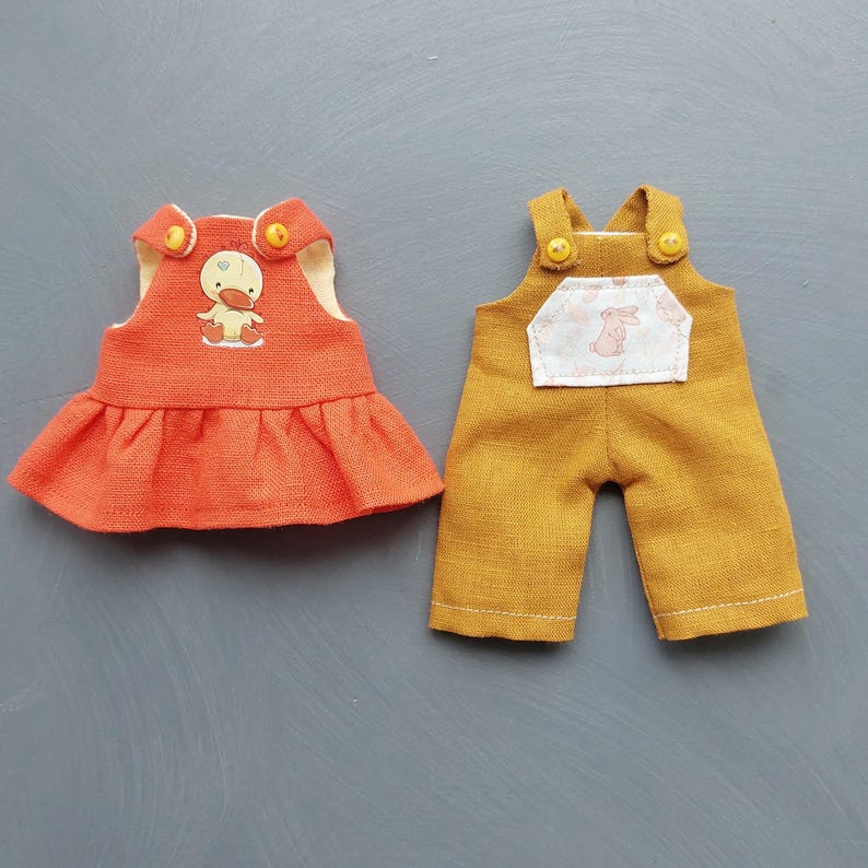 May include: Two doll outfits, one a red dress with a yellow duck applique and the other a mustard yellow overall with a pink bunny applique.
