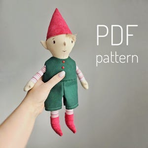 Può includere: Un modello PDF per cucire una bambola di folletto di peluche. Il folletto indossa un cappello rosso, pantaloni verdi e maniche a righe rosse e bianche. La bambola è tenuta in una mano.