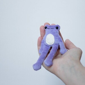 May include: A small purple plush frog with a white belly and pink eyes. The frog is sitting in the palm of a hand.