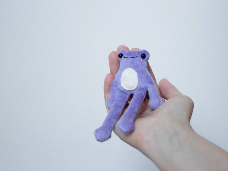 May include: A small purple plush frog with a white belly and pink eyes. The frog is sitting in the palm of a hand.