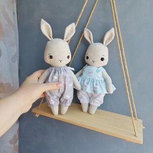 May include: Two handmade bunny dolls with long ears and embroidered faces. The bunny on the left is wearing a light blue and white patterned dress. The bunny on the right is wearing a light blue and white patterned dress with a blue bow. The bunnies are sitting on a wooden shelf.