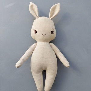 May include: A beige linen bunny rabbit stuffed animal with embroidered eyes and a nose.