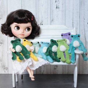 May include: A doll with black hair and a white dress is sitting on a white bench with seven plush frog toys in various shades of green, blue, and purple.