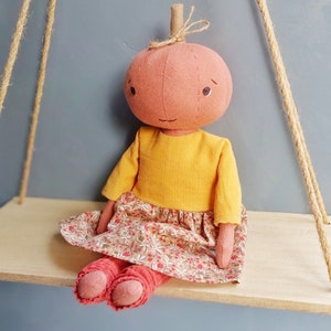 Op de afbeelding: Een handgemaakte pompoendoll met een geel shirt, een rok met bloemenprint en rode leggings. De pop zit op een houten plank.