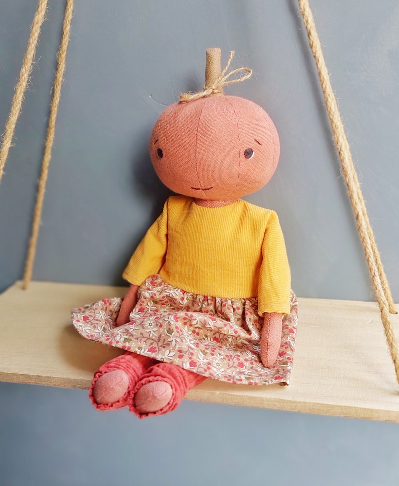Op de afbeelding: Een handgemaakte pompoendoll met een geel shirt, een rok met bloemenprint en rode leggings. De pop zit op een houten plank.