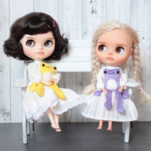 May include: Two dolls with long hair are sitting on a white bench. The doll on the left has black hair and is wearing a white dress with lace trim. She is holding a yellow frog plush toy. The doll on the right has blonde hair and is wearing a white dress with a purple frog plush toy.