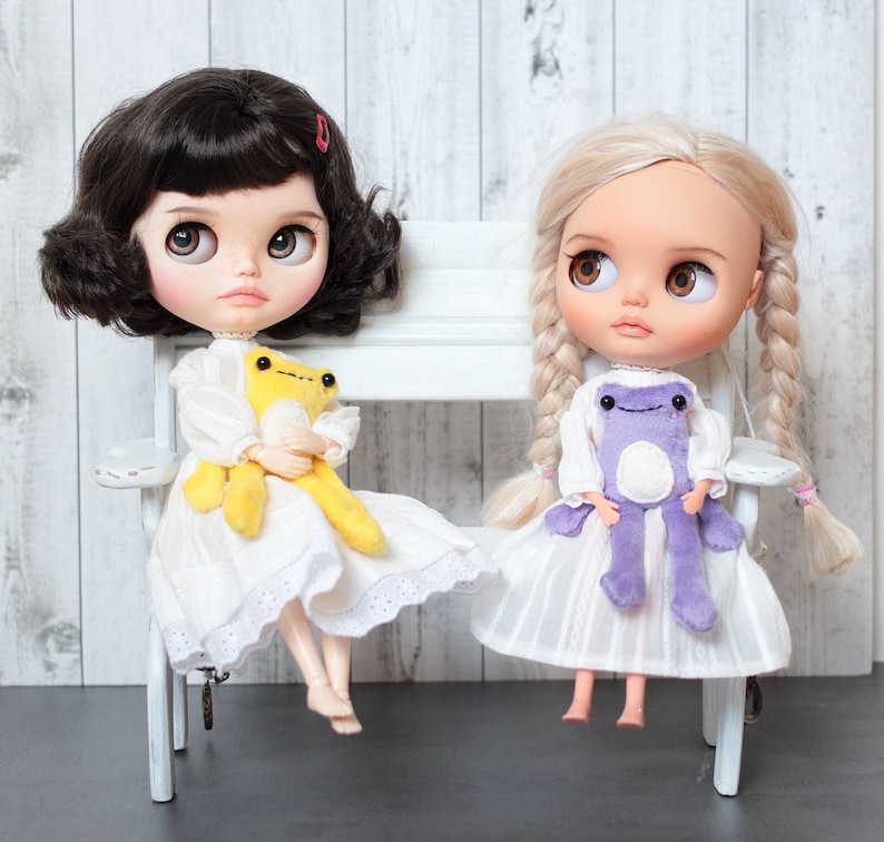 May include: Two dolls with long hair are sitting on a white bench. The doll on the left has black hair and is wearing a white dress with lace trim. She is holding a yellow frog plush toy. The doll on the right has blonde hair and is wearing a white dress with a purple frog plush toy.