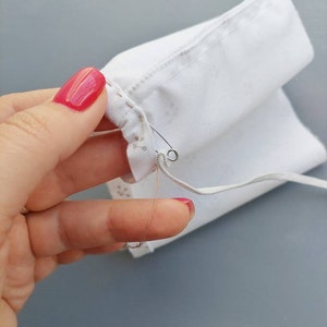 May include: A close-up of a white fabric being sewn with a needle and thread. The fabric is being held in place with a safety pin.