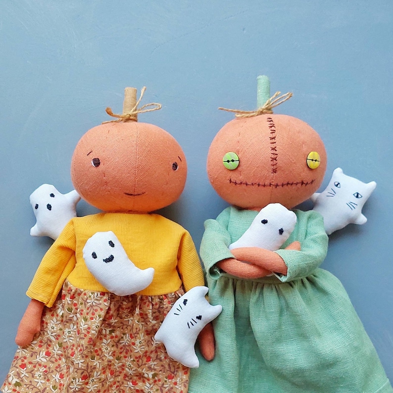 Op de afbeelding: Twee handgemaakte pompoenvormige poppen met gestikte gezichtskenmerken en groene ogen. De poppen houden pluche spook- en katten speelgoed vast. E&eacute;n pop draagt een geel shirt en een bloemenrok, de andere draagt een groene jurk.