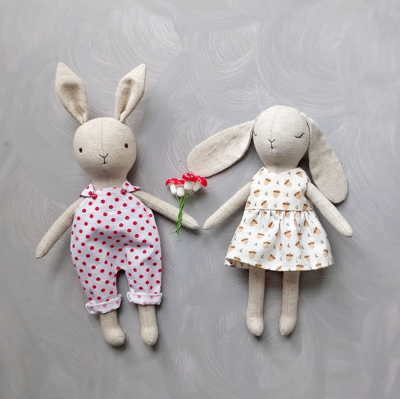 May include: Two handmade plush bunny toys. One bunny is wearing a white dress with a brown acorn pattern. The other bunny is wearing a white and red polka dot overall. Both bunnies have long ears and are made of soft fabric.