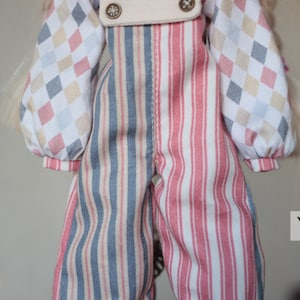 May include: A pair of striped overalls with a blue and white stripe on one leg and a pink and white stripe on the other leg. The overalls have a white bib with two buttons and are made of a soft, cotton fabric.