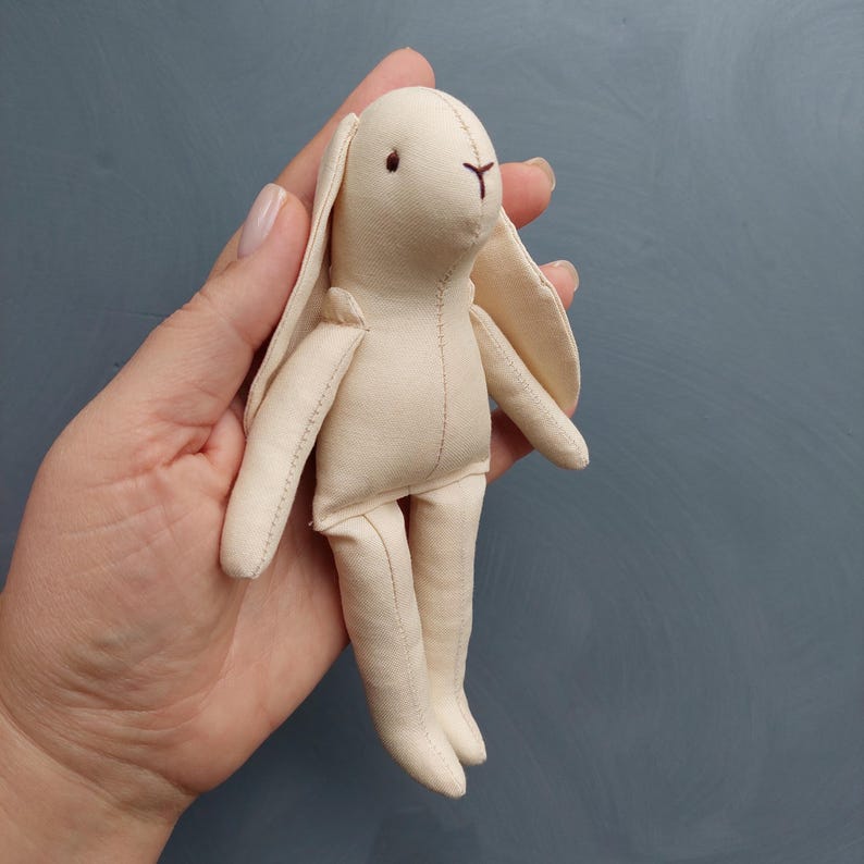 May include: A small, cream-colored fabric bunny with long ears and a stitched face. The bunny is held in a hand.