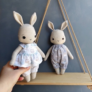 May include: Two handmade fabric bunny dolls with embroidered faces. The doll on the left is wearing a light blue and white dress with an owl print. The doll on the right is wearing a light gray and white dress with a floral print.