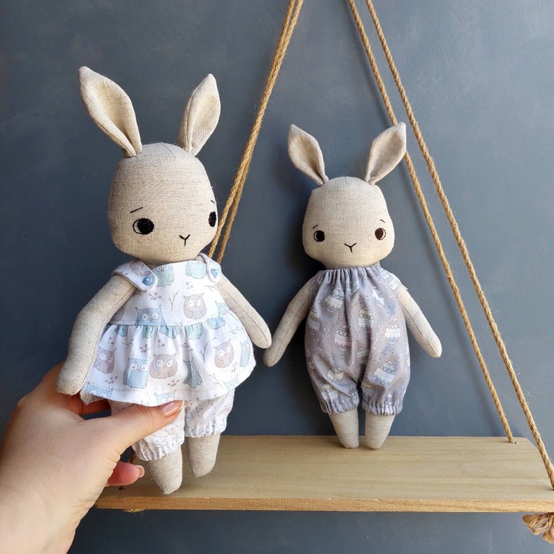 May include: Two handmade fabric bunny dolls with embroidered faces. The doll on the left is wearing a light blue and white dress with an owl print. The doll on the right is wearing a light gray and white dress with a floral print.
