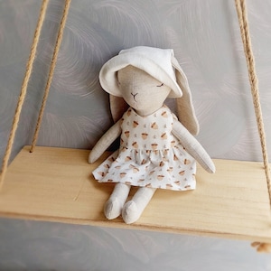 May include: A handmade plush bunny doll wearing a white bonnet and a dress with a brown acorn pattern. The doll is sitting on a wooden shelf.