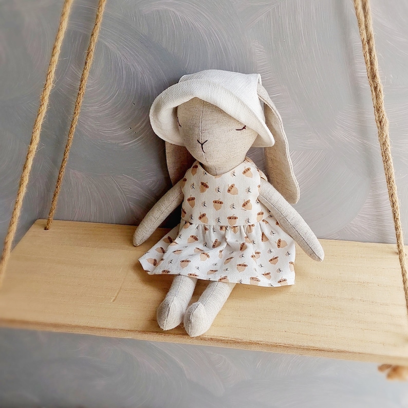 May include: A handmade plush bunny doll wearing a white bonnet and a dress with a brown acorn pattern. The doll is sitting on a wooden shelf.