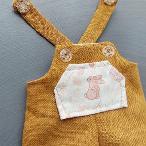 May include: A pair of mustard yellow overalls with a white pocket featuring a pink bunny print. The overalls have adjustable straps with clear plastic buttons.