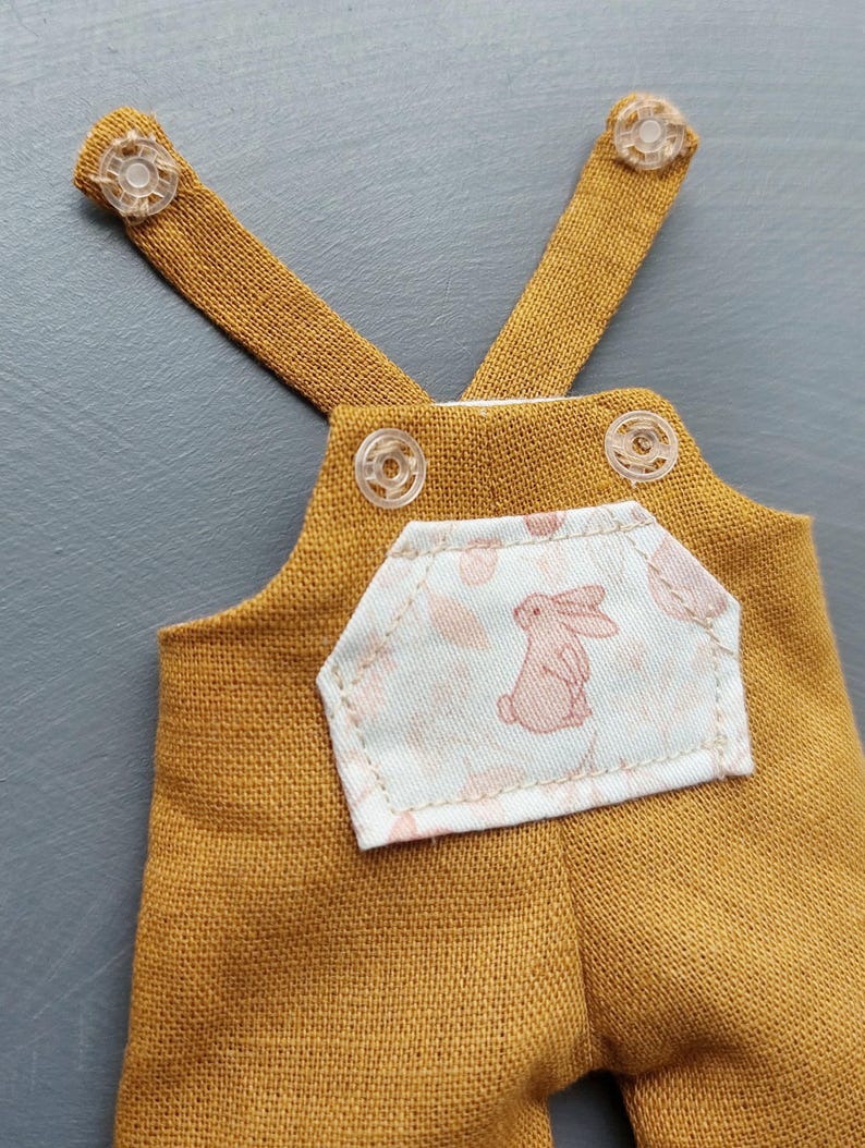 May include: A pair of mustard yellow overalls with a white pocket featuring a pink bunny print. The overalls have adjustable straps with clear plastic buttons.