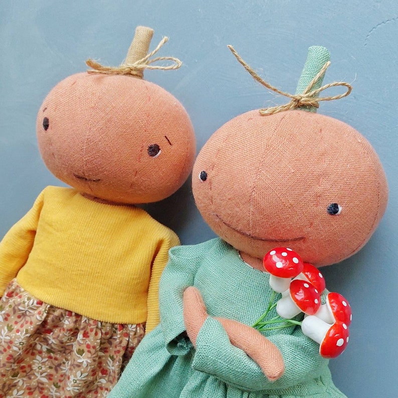Op de afbeelding: Twee handgemaakte stoffen pompoenpoppen. De ene pop draagt een geel shirt en een bloemenrok. De andere pop draagt een groene jurk en houdt een boeket rode en witte paddenstoelen vast.