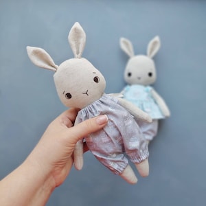 May include: Two handmade plush bunny toys. One is light brown with a gray and white patterned dress and the other is light blue with a white dress. Both have embroidered eyes and noses.