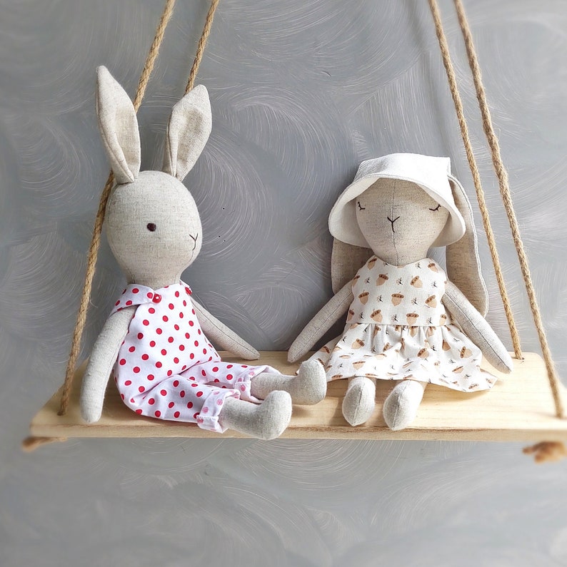 May include: Two plush bunny toys on a wooden swing set. One bunny is wearing a white dress with brown acorn print and a white hat. The other bunny is wearing a red and white polka dot dress.
