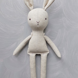 May include: A beige linen bunny rabbit toy with embroidered eyes and a stitched nose. The toy has long arms and legs.