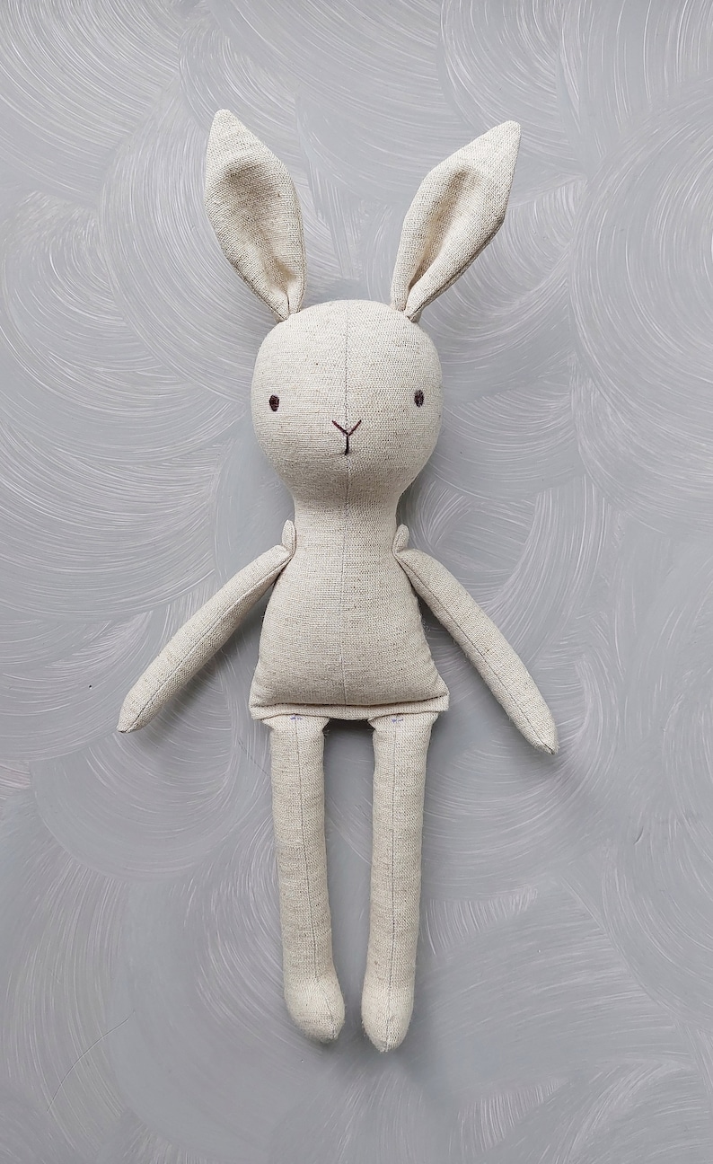 May include: A beige linen bunny rabbit toy with embroidered eyes and a stitched nose. The toy has long arms and legs.