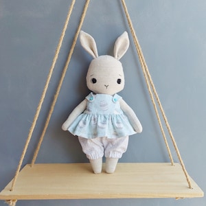 May include: A handmade plush bunny doll wearing a light blue dress with a white and gray egg pattern. The doll is sitting on a wooden shelf with a rope swing hanging above it.