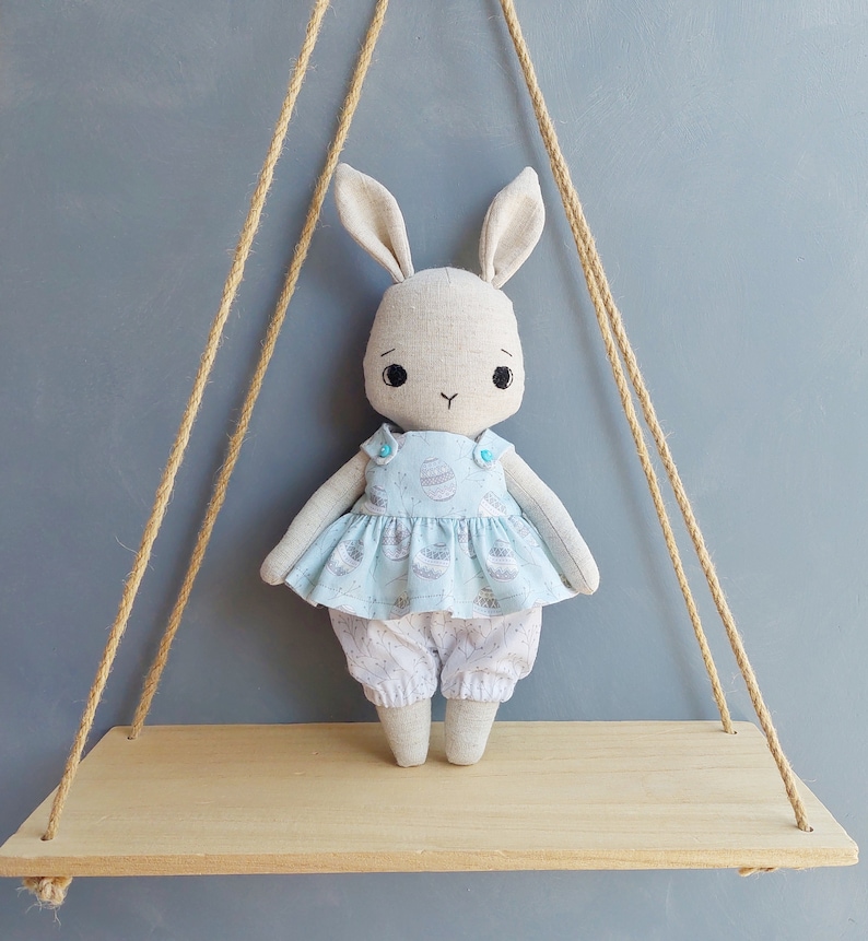 May include: A handmade plush bunny doll wearing a light blue dress with a white and gray egg pattern. The doll is sitting on a wooden shelf with a rope swing hanging above it.