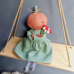 Op de afbeelding: Een handgemaakte pompoendoll die een groene jurk draagt en een boeket rode en witte paddenstoelen vasthoudt. De pop zit op een houten schommel.