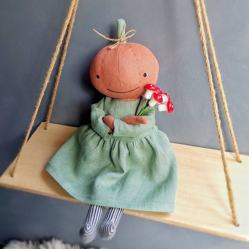 Op de afbeelding: Een handgemaakte pompoendoll die een groene jurk draagt en een boeket rode en witte paddenstoelen vasthoudt. De pop zit op een houten schommel.
