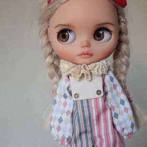May include: A doll with blonde hair in braids wearing a white, pink, and blue checkered shirt with a white collar and a white, pink, and blue striped overall.