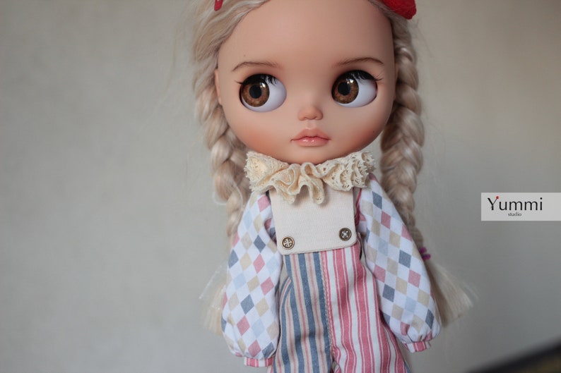 May include: A doll with blonde hair in braids wearing a white, pink, and blue checkered shirt with a white collar and a white, pink, and blue striped overall.