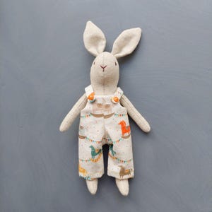 May include: A beige linen bunny toy wearing overalls with a white background and a pattern of colorful horses and flags. The overalls have two orange buttons.