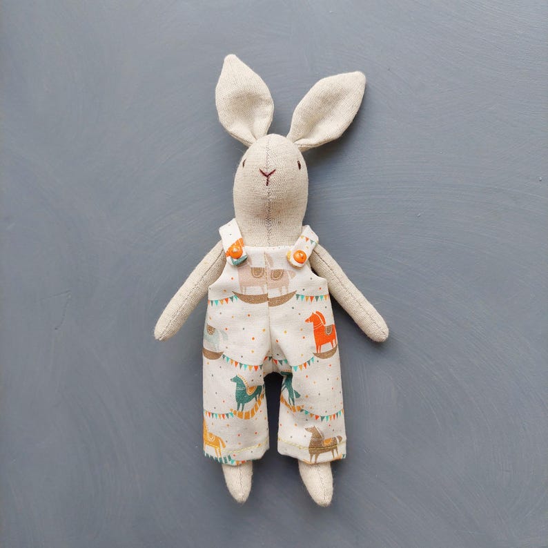 May include: A beige linen bunny toy wearing overalls with a white background and a pattern of colorful horses and flags. The overalls have two orange buttons.