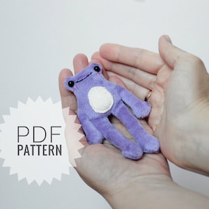 May include: A purple plush frog toy with white eyes and a white belly. The toy is being held in a person's hands. The text "PDF PATTERN" is visible in the background.