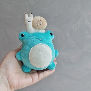 Puede incluir: Una rana de peluche azul con la barriga blanca y un pequeño caracol blanco en la cabeza.