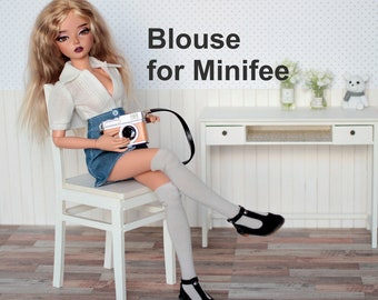 Pode incluir: Padrão PDF para uma blusa para uma boneca Minifee. O padrão é mostrado em uma boneca que usa uma blusa branca, uma saia jeans e meias brancas até o joelho. A boneca está sentada em uma cadeira branca e segurando uma câmera.