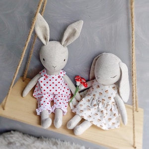 May include: Two handmade plush bunny toys sitting on a wooden shelf. The bunny on the left is wearing a red and white polka dot dress. The bunny on the right is wearing a white dress with a brown acorn pattern.