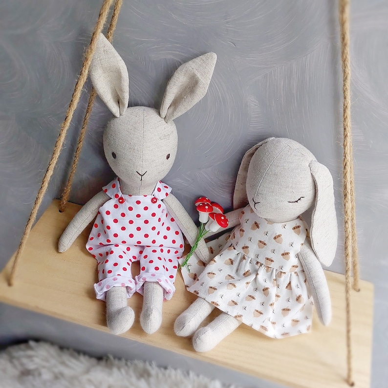 May include: Two handmade plush bunny toys sitting on a wooden shelf. The bunny on the left is wearing a red and white polka dot dress. The bunny on the right is wearing a white dress with a brown acorn pattern.