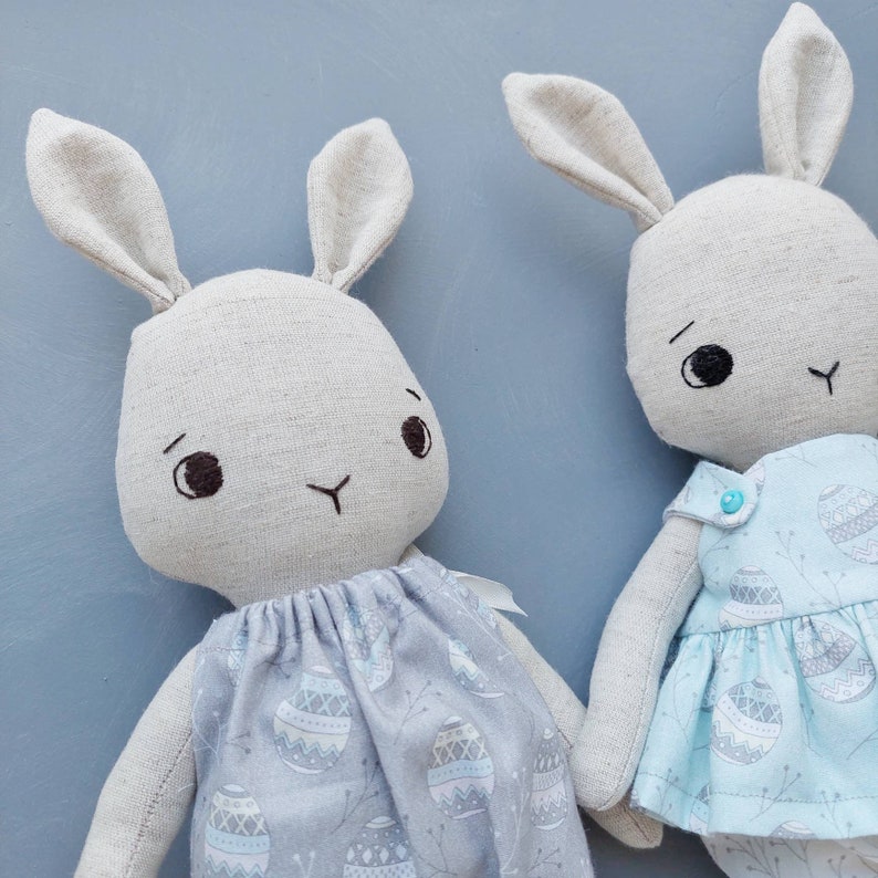 May include: Two handmade plush bunny toys. One bunny is wearing a light blue dress with a white and gray pattern. The other bunny is wearing a gray dress with a white and gray pattern.