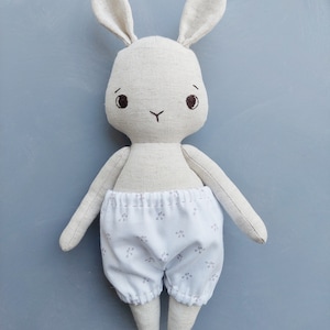 May include: A white linen bunny rabbit toy with embroidered eyes and a stitched nose. The bunny is wearing white cotton pants with a small floral print.
