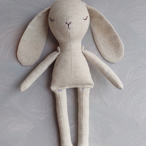 May include: A beige linen bunny rabbit plush toy with embroidered eyes and a stitched nose. The toy has long arms and legs.