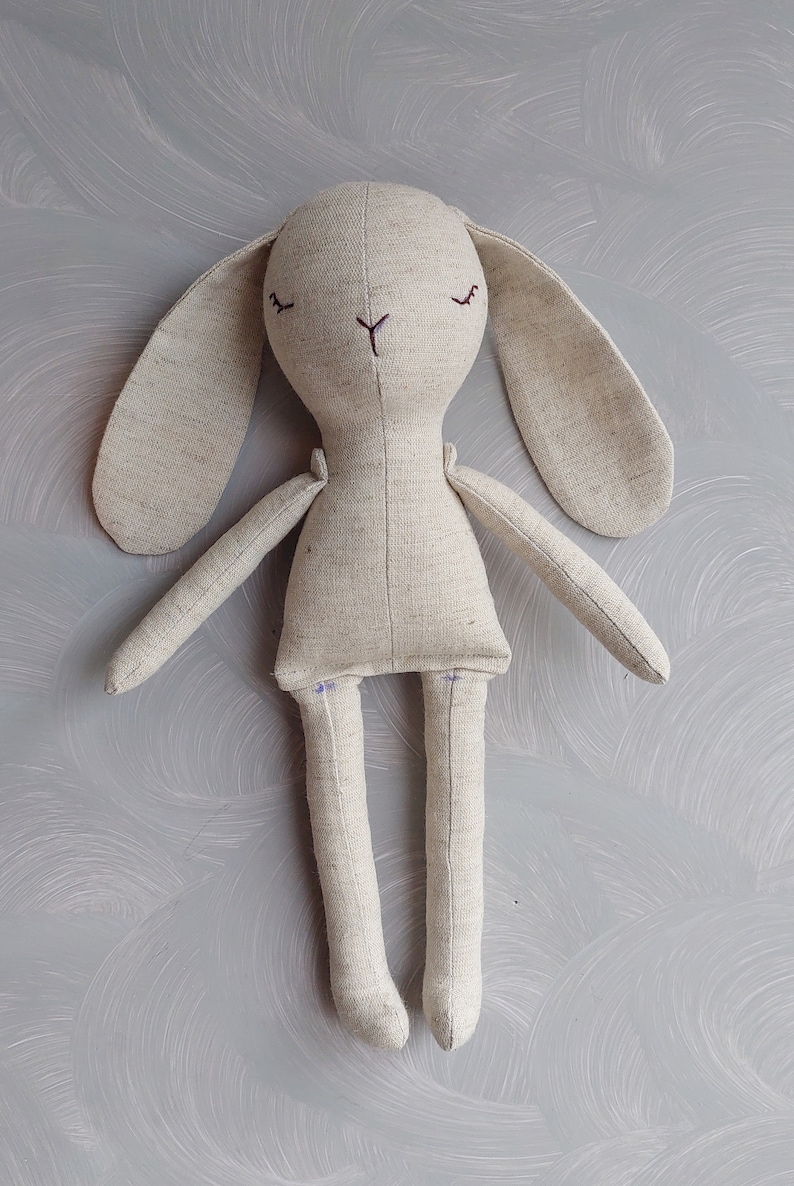May include: A beige linen bunny rabbit plush toy with embroidered eyes and a stitched nose. The toy has long arms and legs.